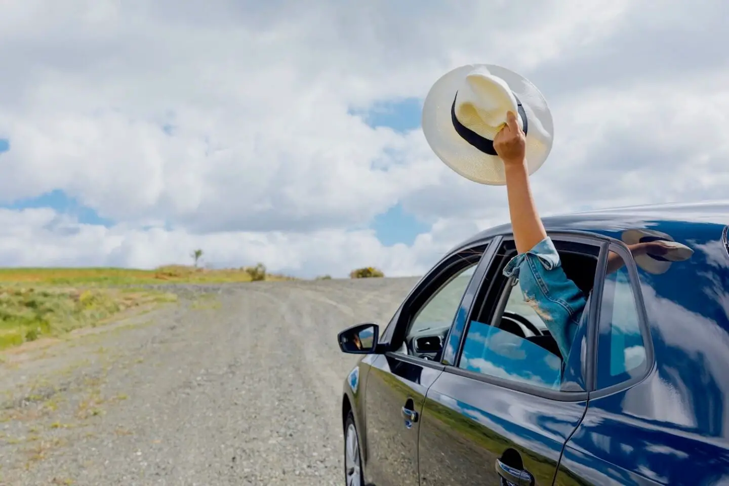 A blue car on a road in the countryside. An arm is coming out of the bar window holding a panama hat