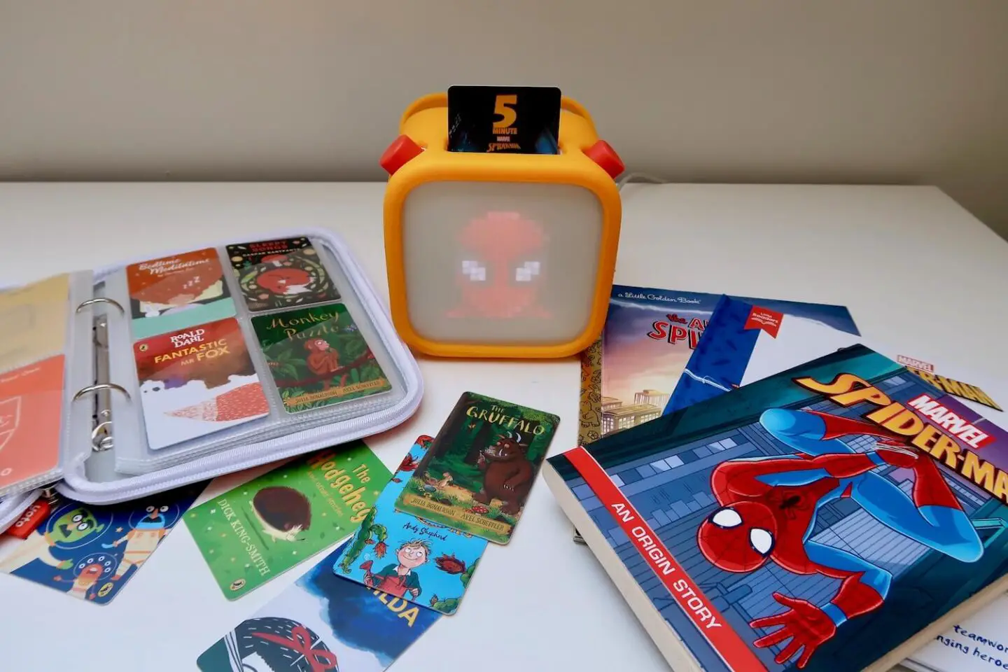 A Yoto player is in the centre of the picture with Spiderman's face is on the display. To the right of the player is a stack of 2 Spiderman books. To the left is a Yoto card holder filled with cards. More cards are scattered in the front.