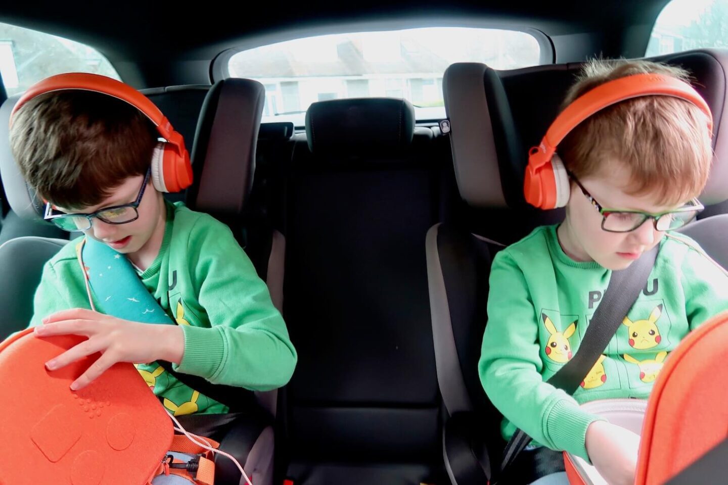 2 children wearing green Pokemon jumpers sitting in the back of the car. They are wearing orange headphones and looking in orange cases on their laps