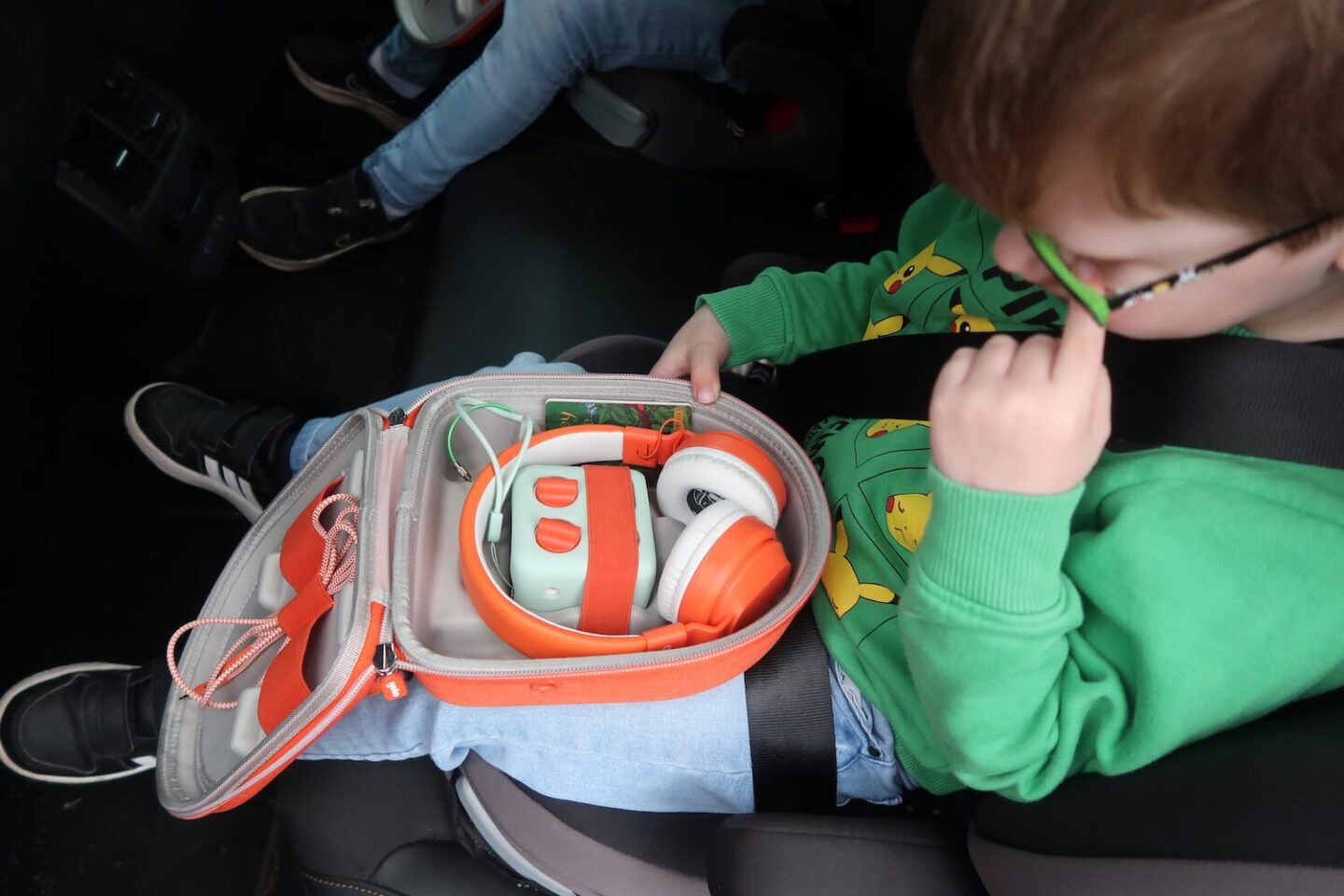 View looking down at a boy in the car. He has a Yoto mini travel case open on his lap with a green Yoto mini and orange headphones in it.