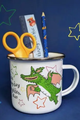 A mug with a picture of a cartoon green dragon on it. A pencil, ruler and scissors are in it