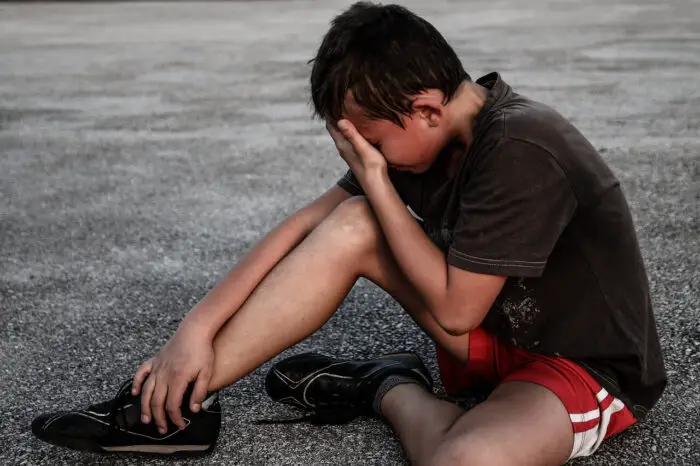 A boy wearing shorts and a t-shirt sitting on the ground. He is covering his face with his hand as if he is crying